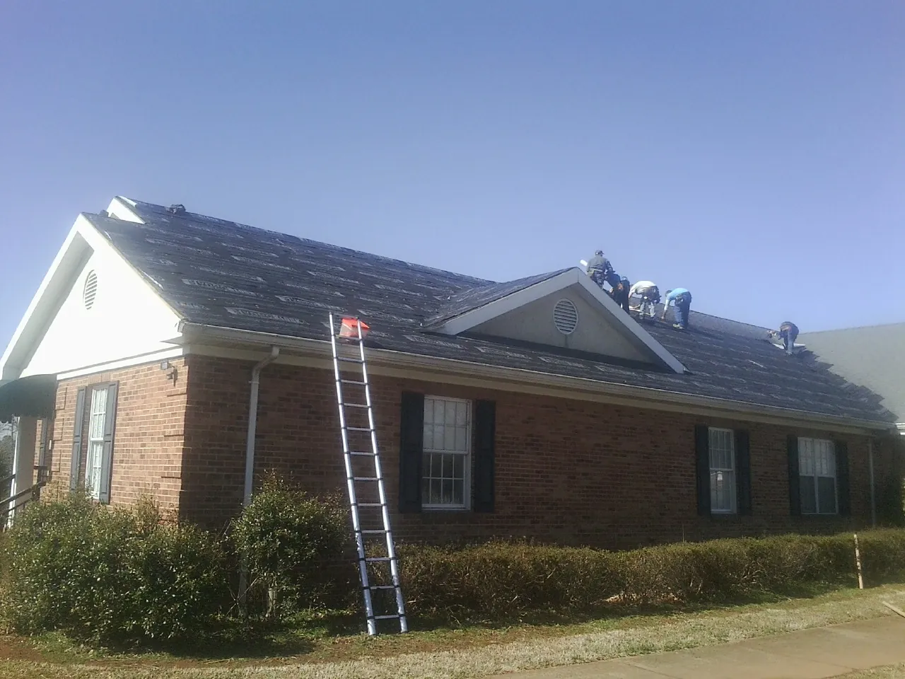 Crew tearing shingles off a brick ranch roof with a ladder set — Emergency Roof Repair work by our crew in Berlin Heights