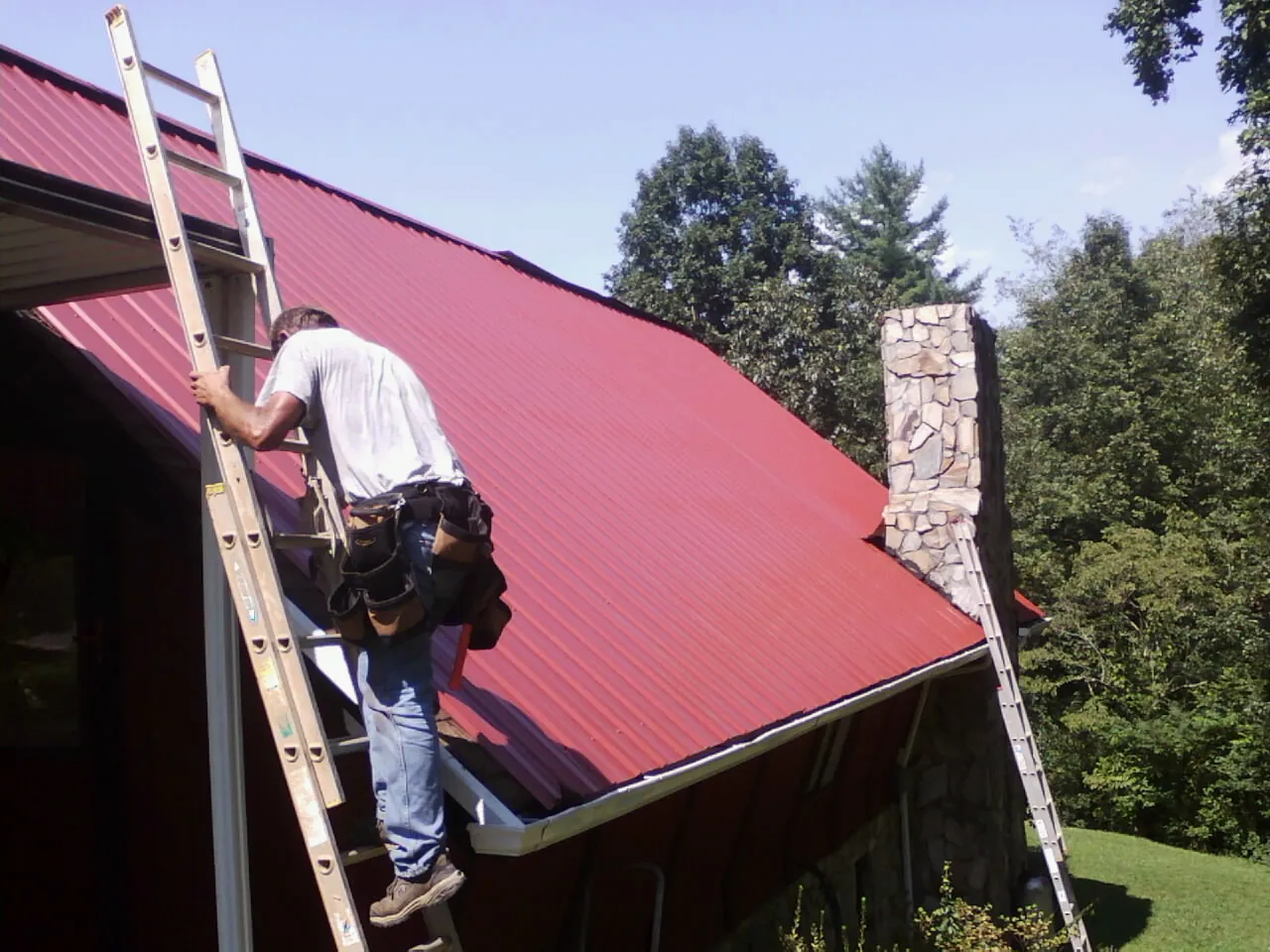 Roofer on a ladder beside a red metal roof and stone chimney — Soffit & Fascia Repair by Roofing Brothers in Berlin Heights